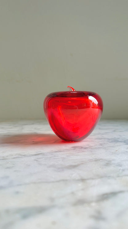 A small, glossy red glass apple-shaped candle or decorative object with a short stem, resting on a light marble surface against a neutral gray background.